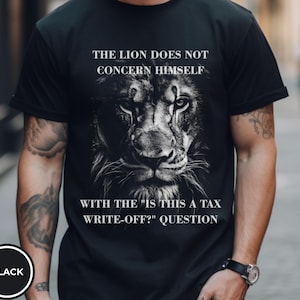 May include: Black t-shirt featuring a detailed black and white image of a lion's face. The text on the shirt reads: "THE LION DOES NOT CONCERN HIMSELF WITH THE 'IS THIS A TAX WRITE-OFF?' QUESTION."