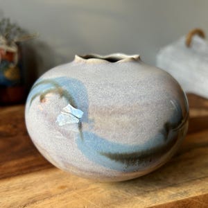 May include: A small, round, ceramic vase with a scalloped rim. The vase is glazed in a light gray color with blue and brown streaks and swirls.