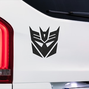May include: Black Transformers logo sticker on a white vehicle. The emblem is a geometric design with sharp angles and a central pointed element, associated with the Transformers franchise. The sticker is applied to the vehicle's body.