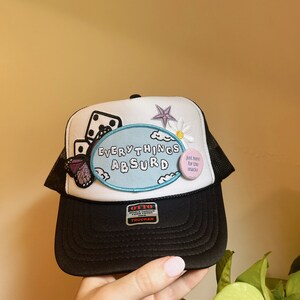 May include: Black trucker hat with a white mesh back. The hat has a blue and white patch that says "EVERYTHING ABSURD" and a pink button that says "Just here for the snacks".