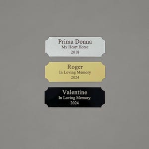 May include: Three engraved metal plaques with rounded corners. The top plaque is silver with the text "Prima Donna My Heart Horse 2018". The middle plaque is gold with the text "Roger In Loving Memory 2024". The bottom plaque is black with the text "Valentine In Loving Memory 2024".