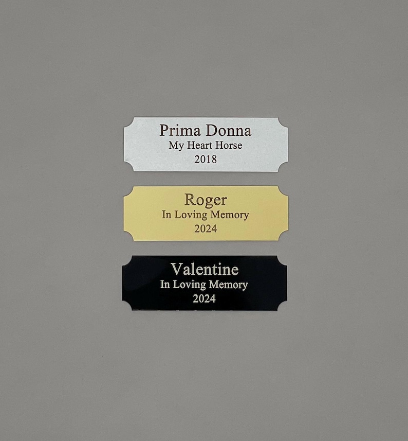 May include: Three engraved metal plaques with rounded corners. The top plaque is silver with the text "Prima Donna My Heart Horse 2018". The middle plaque is gold with the text "Roger In Loving Memory 2024". The bottom plaque is black with the text "Valentine In Loving Memory 2024".