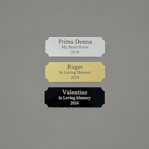 May include: Three engraved metal plaques with rounded corners. The top plaque is silver with the text "Prima Donna My Heart Horse 2018". The middle plaque is gold with the text "Roger In Loving Memory 2024". The bottom plaque is black with the text "Valentine In Loving Memory 2024".