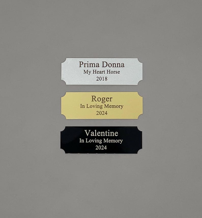 May include: Three engraved metal plaques with rounded corners. The top plaque is silver with the text "Prima Donna My Heart Horse 2018". The middle plaque is gold with the text "Roger In Loving Memory 2024". The bottom plaque is black with the text "Valentine In Loving Memory 2024".