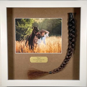 May include: A framed photo of a woman in a white dress and cowboy hat standing with a brown horse in a field of tall grass. A brown horse tail is mounted on the right side of the frame with a gold plaque that reads "Doc" Docs Double straw 001 1997-2024.