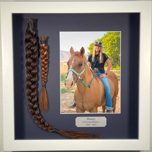 May include: A framed photo of a woman riding a horse with a blue background. The photo is accompanied by two braids of hair, one long and one short, with a plaque that reads "Honey In Loving Memory 1995-2024".
