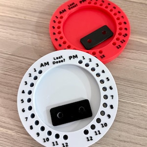 May include: Two circular medication tracking devices, one red and one white, each featuring a central recessed area with a black rectangular slider. The outer rim is printed with the text "Last Dose?" and "AM" and "PM" labels, alongside numbered holes from 1 to 12 for both time periods to track daily medication intake.