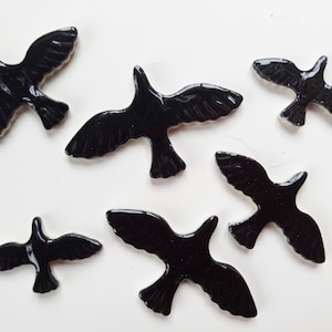 May include: Six glossy black bird-shaped embellishments are scattered on a white surface. The birds are in various positions, with wings spread in flight. The smooth, reflective surface of the birds catches the light, creating highlights.