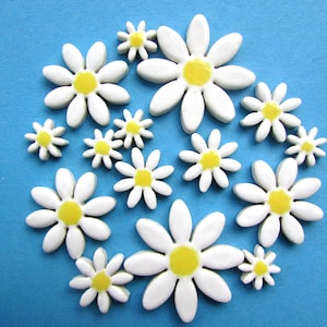 Puede incluir: Un conjunto de adornos blancos con forma de margarita y centros amarillos. Las margaritas, de varios tamaños, están dispuestas sobre un fondo azul. Los pétalos lisos crean un efecto alegre y decorativo.