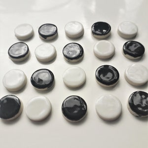Può includere: Una collezione di venti piccoli pezzi rotondi in ceramica. Ogni pezzo è bianco con una parte superiore smaltata nera o nero con una parte superiore smaltata bianca. I pezzi sono disposti in una griglia, creando un contrasto visivo accattivante.