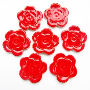 May include: Seven glossy, red ceramic flower-shaped embellishments arranged on a white background. Each flower has multiple petals and a central indentation, creating a three-dimensional effect. The vibrant red color and smooth surface suggest a decorative or craft item.