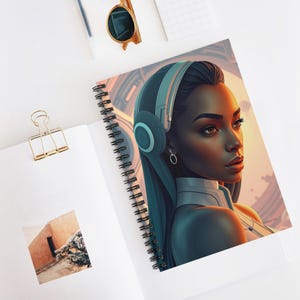 Puede incluir: Cuaderno de espiral con un retrato de arte digital de una mujer de piel oscura con auriculares. La obra de arte tiene un tema futurista con un fondo cálido, naranja y amarillo. El cuaderno está abierto, revelando la obra de arte y una página en blanco.