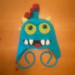 May include: A blue crocheted monster hat with yellow horns, green eyes, white teeth, and a red tongue. The hat has a long blue tie that can be tied under the chin.