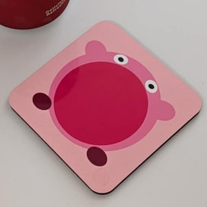 May include: A red Coca-Cola can beside a pink square coaster with a cartoon character design. The coaster has a light pink background, a darker pink centre, and a cartoon face.
