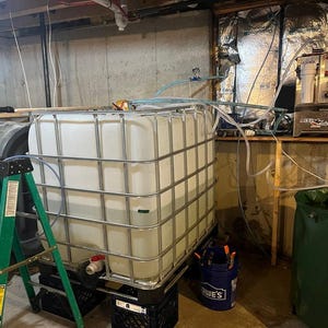 May include: Two large white plastic storage tanks with metal cages, one on a black pallet and the other on a green step ladder. A silver water filtration system is on a wooden shelf in the background.