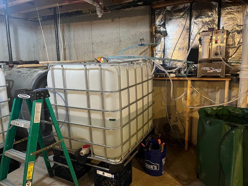 May include: Two large white plastic storage tanks with metal cages, one on a black pallet and the other on a green step ladder. A silver water filtration system is on a wooden shelf in the background.