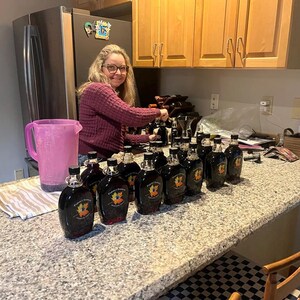 May include: A woman in a purple sweater is filling glass bottles with a dark liquid. The bottles are lined up on a granite countertop. The bottles have a label with a maple leaf design.