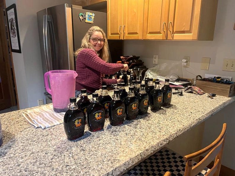 May include: A woman in a purple sweater is filling glass bottles with a dark liquid. The bottles are lined up on a granite countertop. The bottles have a label with a maple leaf design.