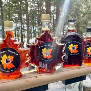 May include: Four glass maple syrup bottles shaped like maple leaves. Each bottle has a label with the text "Pishou's Sucrerie" and a picture of a maple leaf. The bottles are on a wooden surface.
