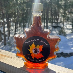 May include: A maple syrup bottle shaped like a maple leaf with a black label that says "Pishou's Sucrerie, Chesterville, Maine".