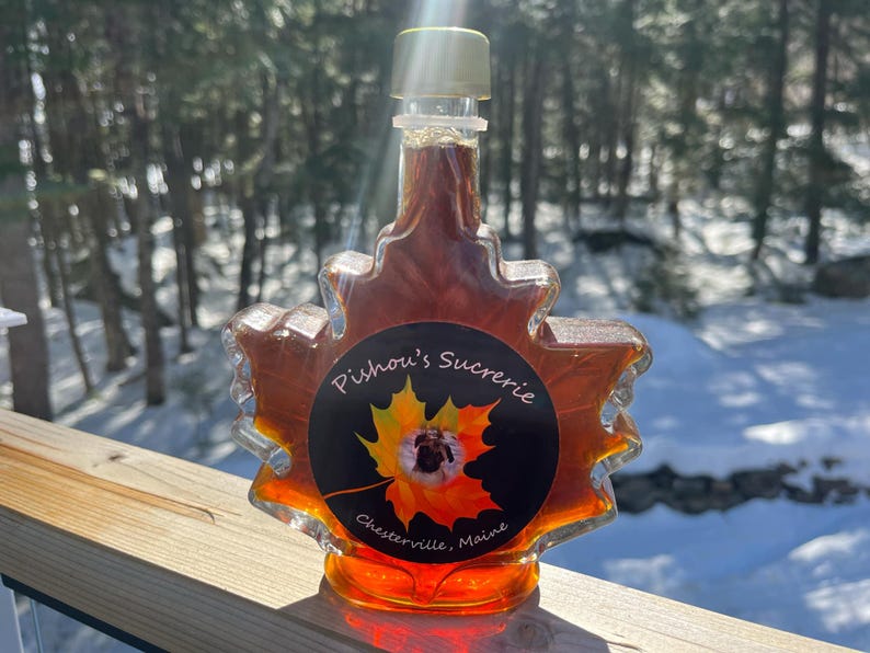 May include: A maple syrup bottle shaped like a maple leaf with a black label that says "Pishou's Sucrerie, Chesterville, Maine".