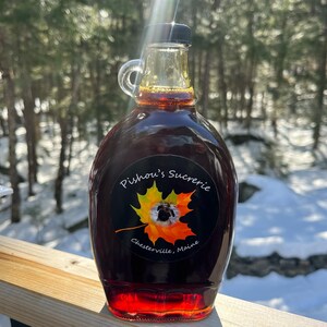 May include: A glass bottle of maple syrup with a black label that reads "Pishou's Sucrerie, Chester ville, Maine". The label features a yellow and orange maple leaf with a small black dog inside.