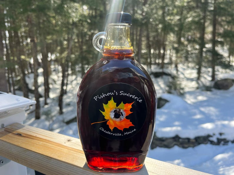 May include: A glass bottle of maple syrup with a black label that reads "Pishou's Sucrerie, Chester ville, Maine". The label features a yellow and orange maple leaf with a small black dog inside.