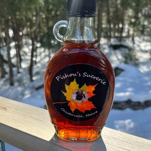 May include: A glass bottle of maple syrup with a black label that reads "Pishou's Sucrerie, Chester ville, Maine". The label features a stylized maple leaf with a bee inside.