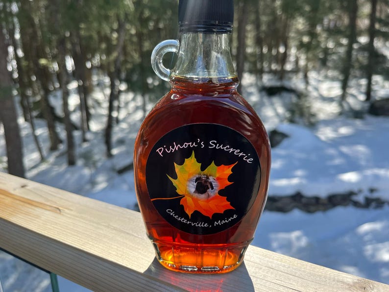 May include: A glass bottle of maple syrup with a black label that reads "Pishou's Sucrerie, Chester ville, Maine". The label features a stylized maple leaf with a bee inside.