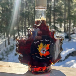 May include: A glass bottle of maple syrup in the shape of a maple leaf. The bottle is filled with dark amber syrup and has a label that reads "Pishon's Sucrerie, Chesterville, Maine".