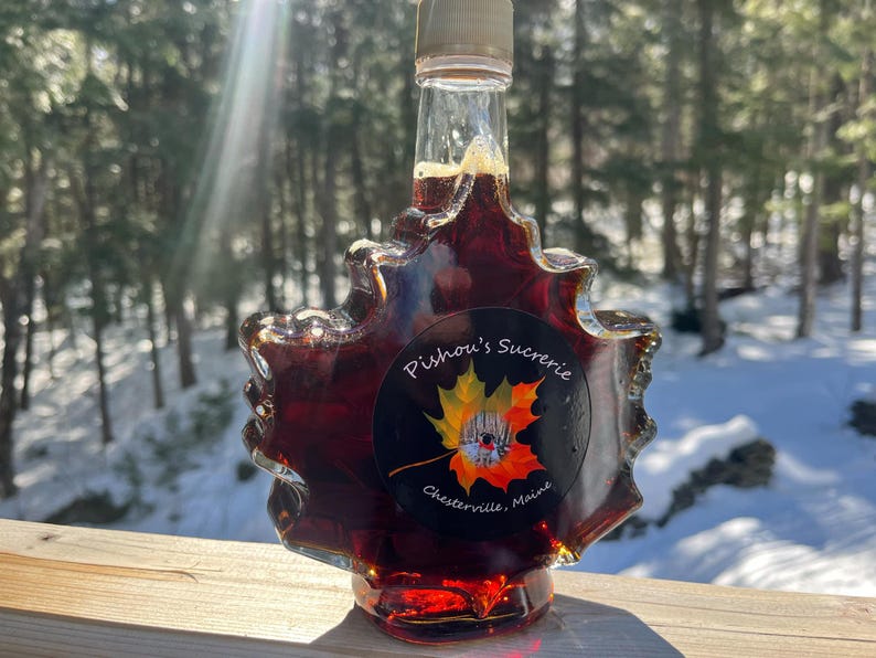 May include: A glass bottle of maple syrup in the shape of a maple leaf. The bottle is filled with dark amber syrup and has a label that reads "Pishon's Sucrerie, Chesterville, Maine".