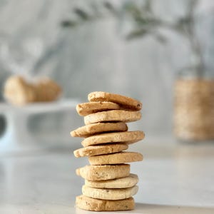 May include: A stack of golden-brown, round cookies with visible specks, arranged in a tower. The cookies are on a white surface, with a blurred background featuring a vase and a decorative item. The cookies appear to be homemade.