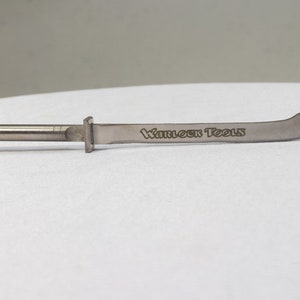 Może przedstawiać: Srebrne metalowe narzędzie z zakrzywionym ostrzem i uchwytem. Ostrze jest grawerowane tekstem "Warlock Tools".
