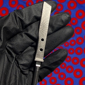 May include: A stainless steel knife with a decorative pattern of circles and dots. The knife has a long, rectangular blade with three circular holes and a textured handle. The blade's design features concentric circles and a stylized sunburst pattern.