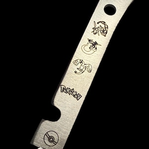 May include: A stainless steel bottle opener with a curved blade and a circular hole. The opener features engraved Pokémon characters and the word "Pokémon". A Poké Ball symbol is also engraved on the opener.