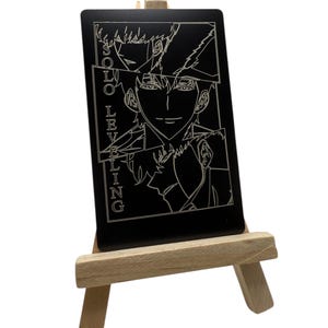 May include: A black card with a white illustration of a character from the Solo Leveling series, displayed on a small wooden easel. The card features the text "SOLO LEVELING" vertically on the left side.