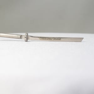 Könnte beinhalten: Ein silberfarbenes Metallwerkzeug in Form eines Miniatur-Katanas. Das Werkzeug hat ein Gewindeende, einen kreisförmigen Schutz und eine Klinge mit dem eingravierten Text "WARLOCK TOOLS". Das Werkzeug liegt auf einer weißen Oberfläche.