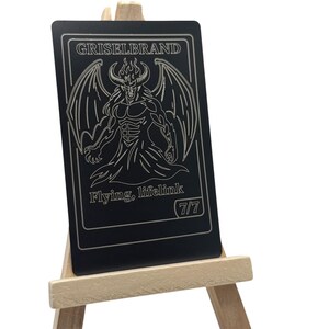 Puede incluir: Una tarjeta negra con la palabra "Griselbrand" en la parte superior y una ilustración detallada de un demonio alado. El texto "Flying, lifelink" está debajo, con "7/7" en un círculo. La tarjeta se muestra en un pequeño caballete de madera.