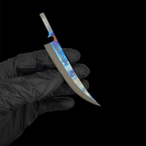 May include: A small, curved knife with a textured handle and a blade featuring a colorful, marbled pattern of blues, oranges, and purples. The text "Dabbin Aladdin" is visible on the blade. The knife is held against a black background.
