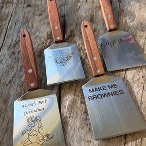 May include: Four stainless steel spatulas with wooden handles. Each spatula has a different engraved design. The designs include a skull with wings, the text "World's Best Grandma" with a cartoon donkey, the text "Make Me Brownies", and the text "Chef Nat".