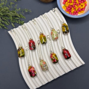 Puede incluir: Un conjunto de ocho dijes en forma de frascos de vidrio en miniatura, rellenos con alimentos de arcilla polimérica colorida como pepinillos, verduras y frutas. Cada frasco está unido a una cadena de metal plateado con un cierre de mosquetón.