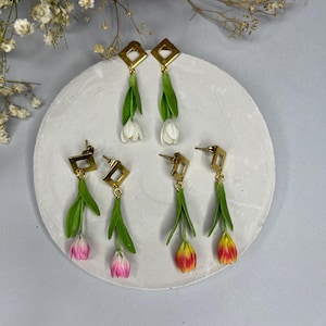 Op de afbeelding: Oorbellen met tulpontwerpen in wit, roze en geel. Elke oorbel heeft een goudkleurige vierkante bovenkant, een groene stengel en een tulpbloem. De oorbellen worden gepresenteerd op een wit cirkelvormig oppervlak.
