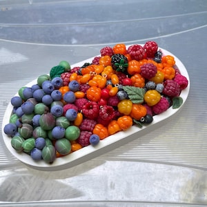 Op de afbeelding: Een wit ovaal dienblad gevuld met een verzameling miniatuur kunstbessen en vruchten. Het assortiment bevat blauwe bosbessen, groene kruisbessen, oranje kruipbramen, rode frambozen en een enkele zwarte braam met groene blaadjes.