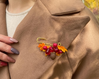May include: A gold-toned safety pin brooch featuring a cluster of small orange and yellow fabric leaves, several glossy red spherical beads, and a single brown acorn charm attached to the metal pin base, pinned to a tan wool coat.