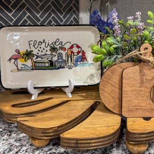 May include: A wooden tray with a carved leaf design, featuring a white ceramic tray with a black border and the word "Florida" in black script. The ceramic tray has a colorful illustration of Florida landmarks and a pink flamingo. The tray is also decorated with two wooden cutting boards, one with a brown finish and the other with a natural wood finish. The cutting board with the brown finish has the letter "C" carved into it.