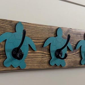 May include: A rectangular wooden wall rack featuring three teal-colored turtle cutouts mounted on a dark stained wood plank. Each turtle has a black metal double hook attached to its center, providing functional storage for hanging various items.