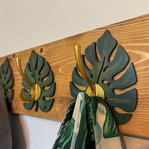 May include: Wooden wall-mounted coat rack with three hooks. The rack features dark green monstera leaf cutouts and gold-colored hooks. The wooden board has a natural wood grain finish. A gray garment and a green and white patterned scarf hang from the hooks.