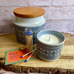 May include: A white ceramic candle jar with a blue and green floral pattern. The jar has a wooden lid and is filled with a white candle. A small glass jar with matches is next to the candle jar. A small card with the text "Bee Loves Bizarre" is in the foreground.
