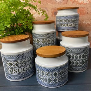 May include: A set of white ceramic kitchen storage jars with wooden lids. The jars have a blue and green geometric design and are labelled "SUGAR" and "COFFEE". They are arranged on a dark blue surface.