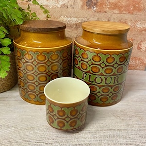 May include: A collection of three ceramic kitchen storage jars with wooden lids. The jars are in shades of brown and green with a retro design. One jar has the word "FLOUR" in green. A small matching cup is in front.